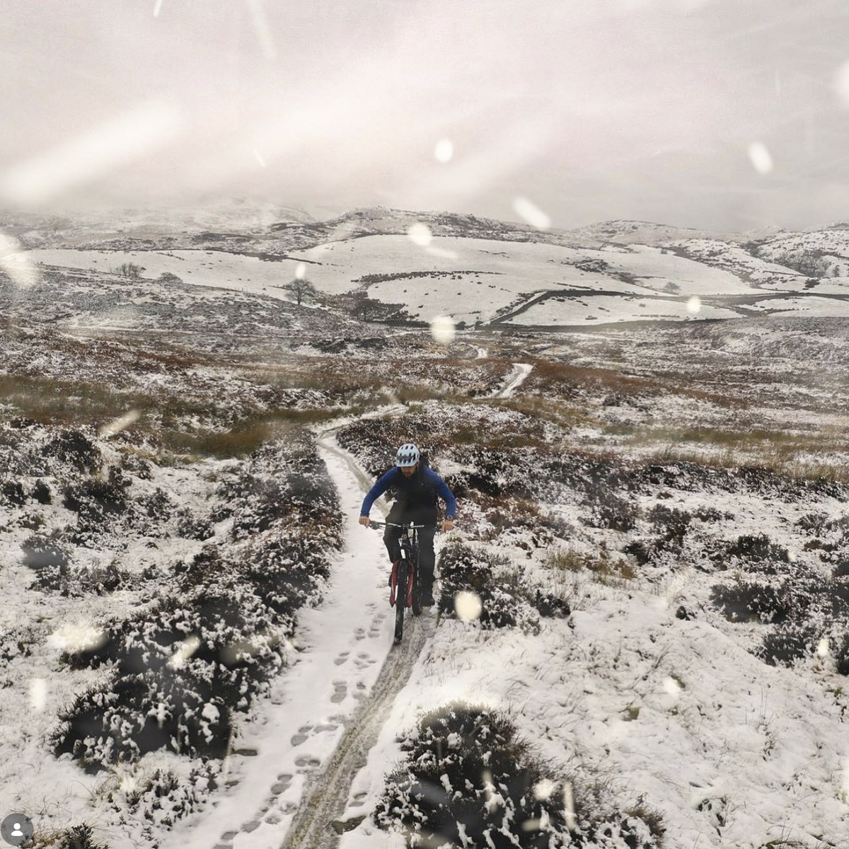 Mountain biker riding a snow-covered trail across open moorland during a snowfall