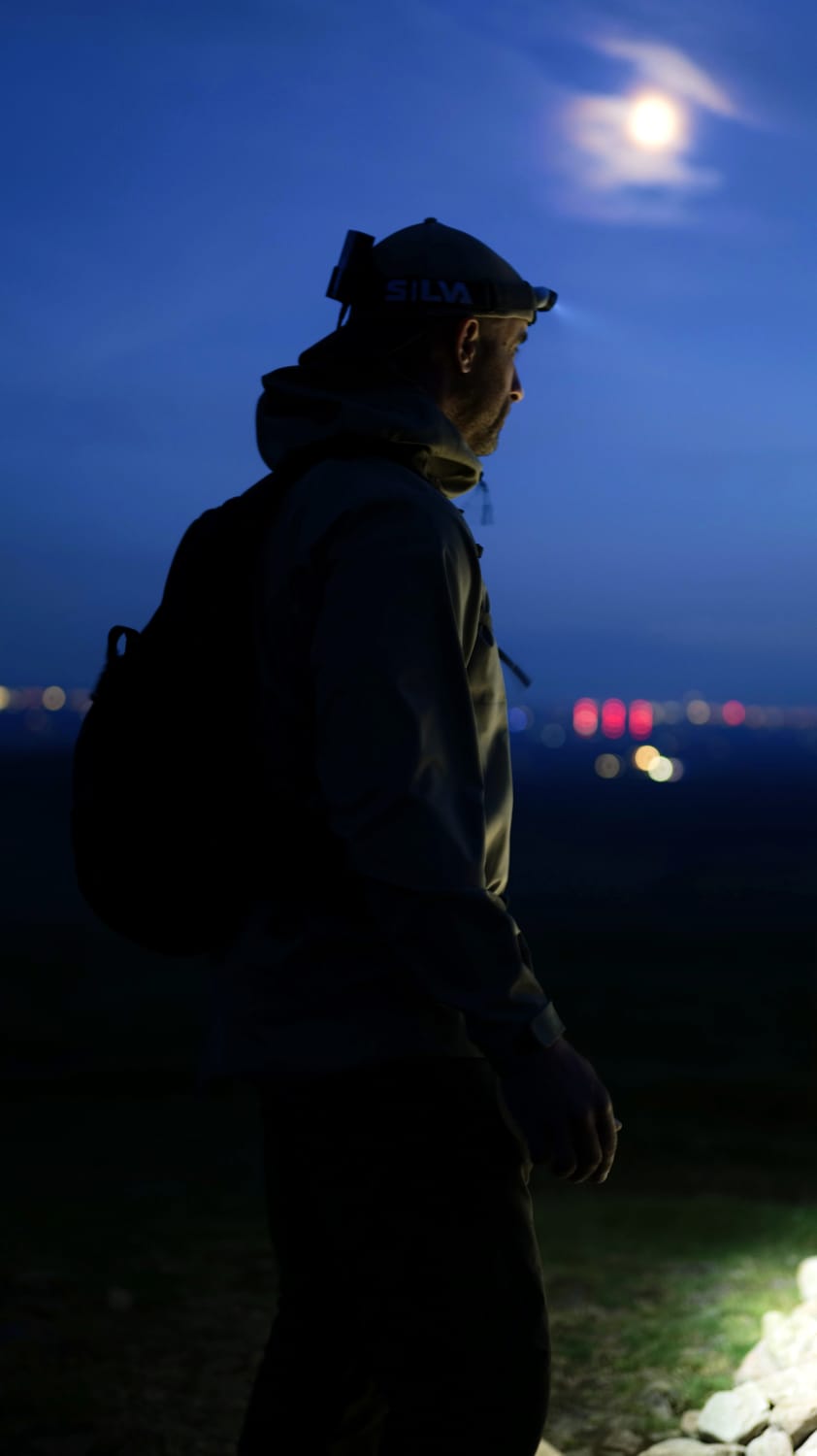 Silhouette of a hiker wearing a headlamp and backpack at dusk, standing under a full moon with distant city lights below