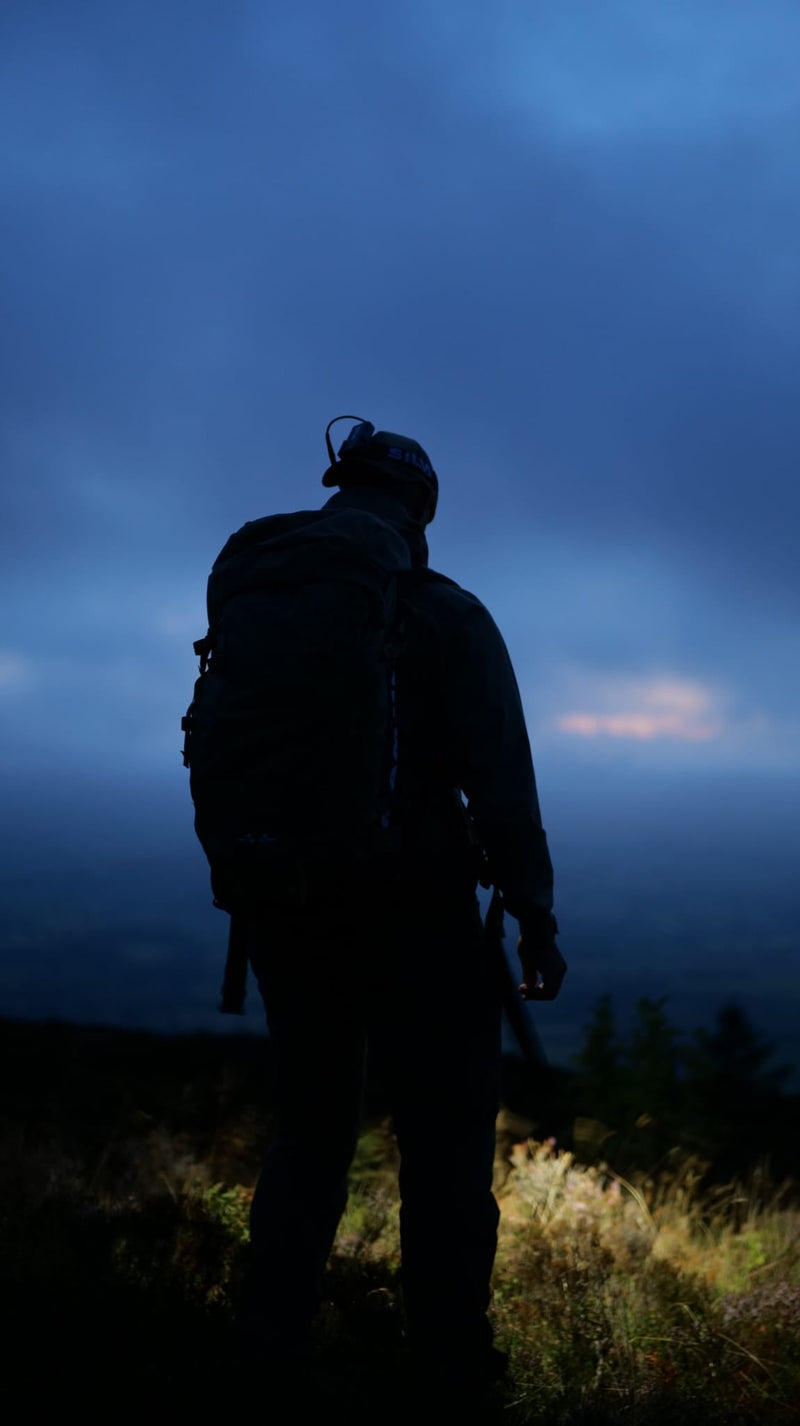 Silhouette of a hiker with a backpack and headlamp standing on a hillside overlooking a misty valley at dusk
