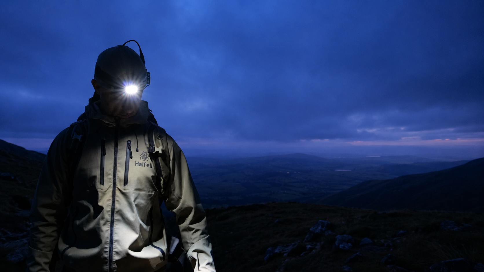 Man wearing a Halfell hardshell jacket and headlamp on a hillside at dusk, overlooking a wide valley
