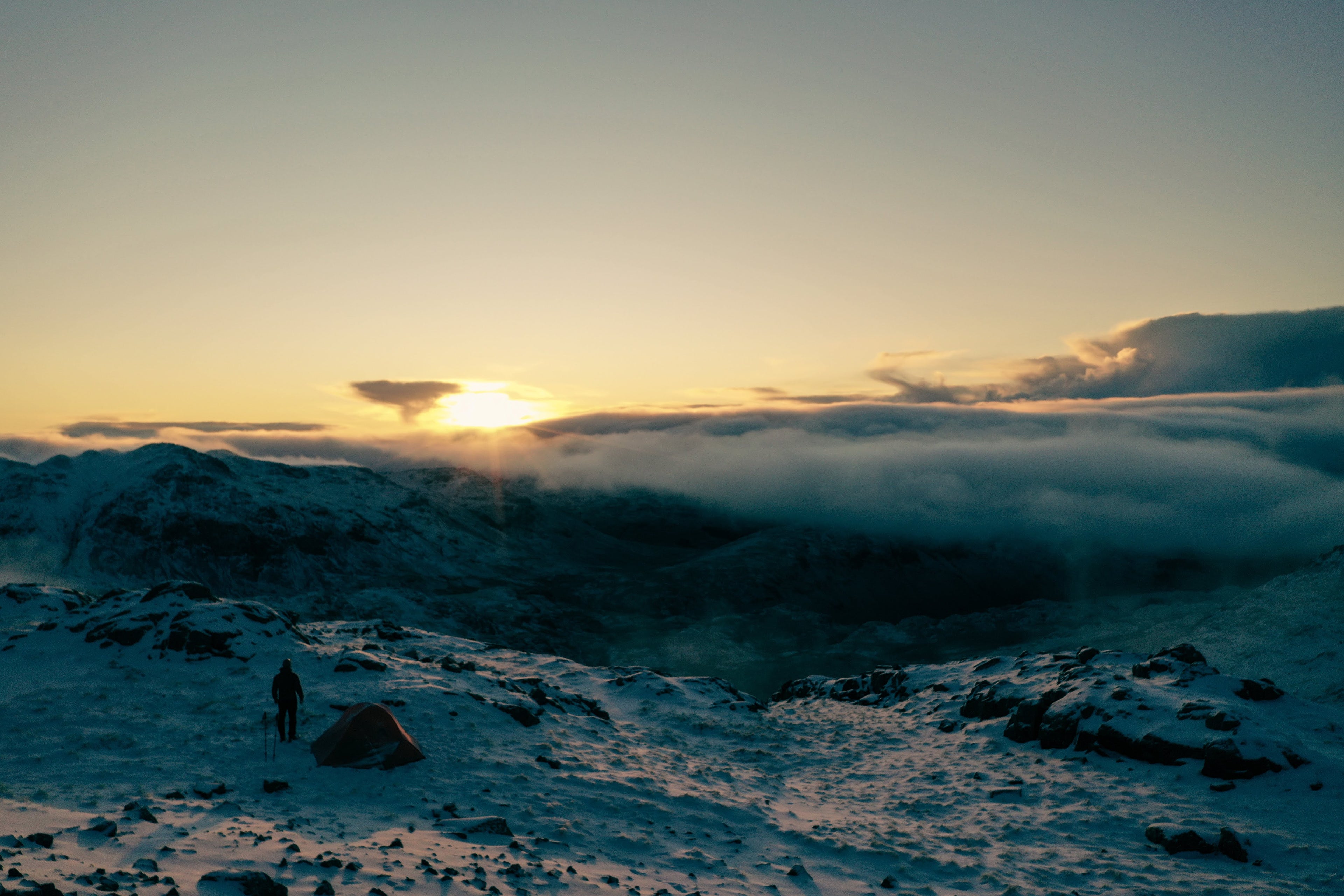 Snow-covered mountain ridge at sunrise with a hiker and tent above a cloud inversion filling the valley below